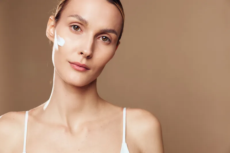 Young woman with natural skin applying white face cream on cheek, looking at camera, isolated on beige background. Concept of skincare, beauty, wellness, and self-care routine.