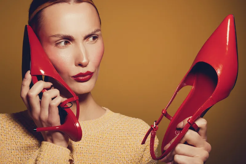 Woman with red lipstick holds red high heel shoes near face, looking thoughtfully to side. Yellow background and beige sweater create warm, stylish fashion concept with focus on footwear.