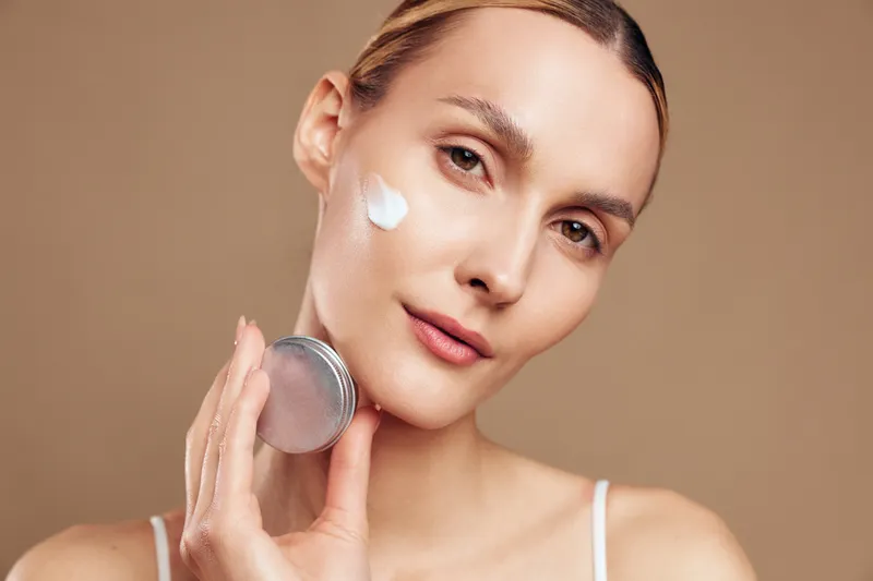 Young woman applies moisturizing cream on her face with gentle smile on beige background skincare routine beauty concept closeup portrait of female model with clear skin.