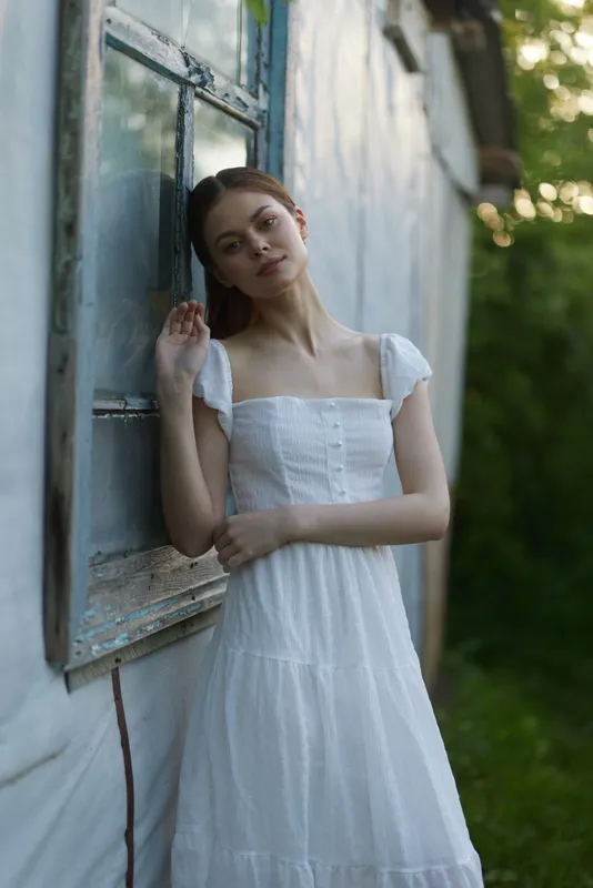 Young woman in a white dress standing by a window, conveying serenity and natural beauty in a green outdoor setting, perfect for promoting relaxation and summer vibes