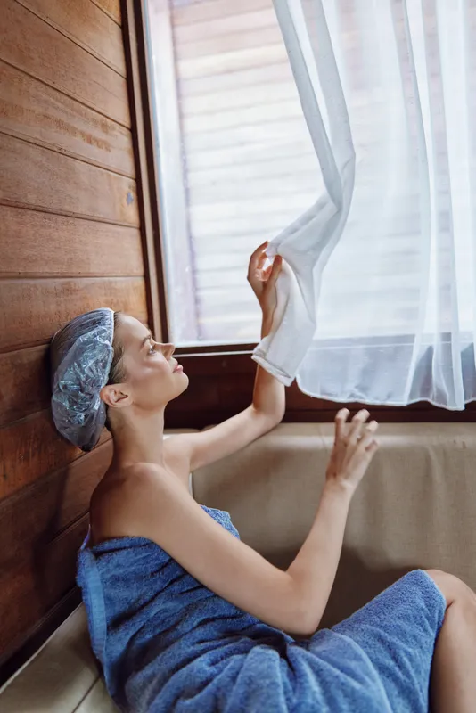 young woman wrapped in blue towel and shower cap relaxing indoors near wooden wall and sheer white curtain in serene spa setting with soft natural light