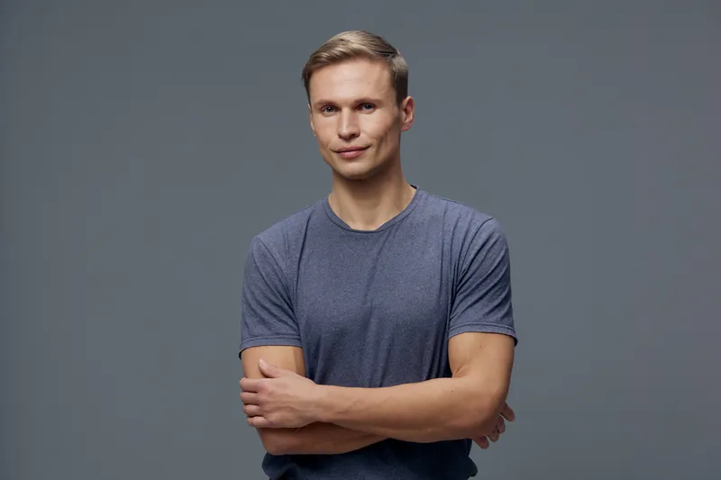 Confident young man with short blond hair wearing casual blue t-shirt standing with arms crossed isolated on plain gray background studio portrait. People concept