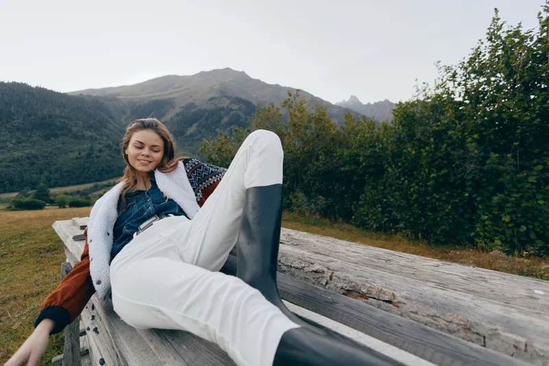 Woman smile mountains outdoors bench boots relaxed nature portrait of a young woman lounging on a wooden bench near alpine hills, casual jacket and white pants in serene countryside scene
