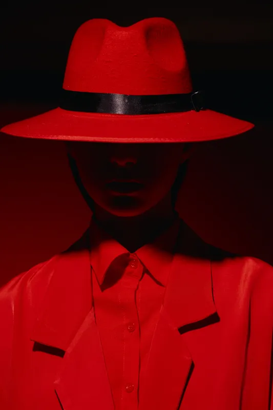 Mysterious woman in a red fedora hat and matching coat poses with shadowed face on red background. High fashion beauty portrait showcasing creative bold style.