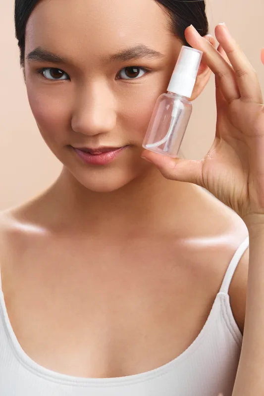 Asian woman holds a small spray bottle near her face, showcasing skincare product in a soft, clean setting. She smiles gently, conveying confidence and self care.