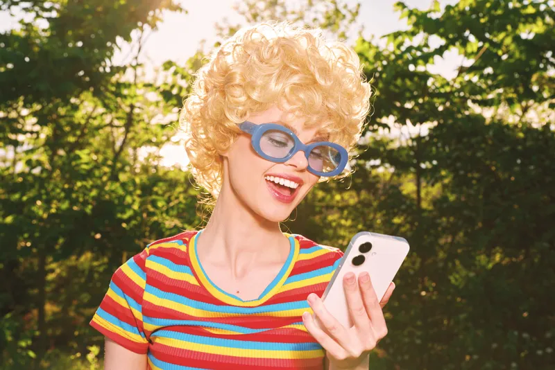 Cheerful woman outdoors, retro vibe, striped colorful shirt, curly wig, blue glasses, smiling at smartphone, sunlit park, candid moment, warm tones, natural greenery backdrop glow