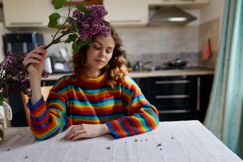 Woman sits at a table in a bright kitchen, holding lilac flowers, wearing a striped sweater, relaxed mood, candid lifestyle shot, natural light, cozy home atmosphere, indoors.
