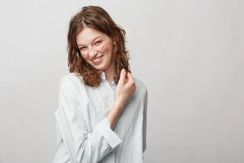 Smiling young woman with curly hair wearing white shirt looks playful and happy. Studio portrait on light background. Concept of joy, youth, casual style, and natural beauty.