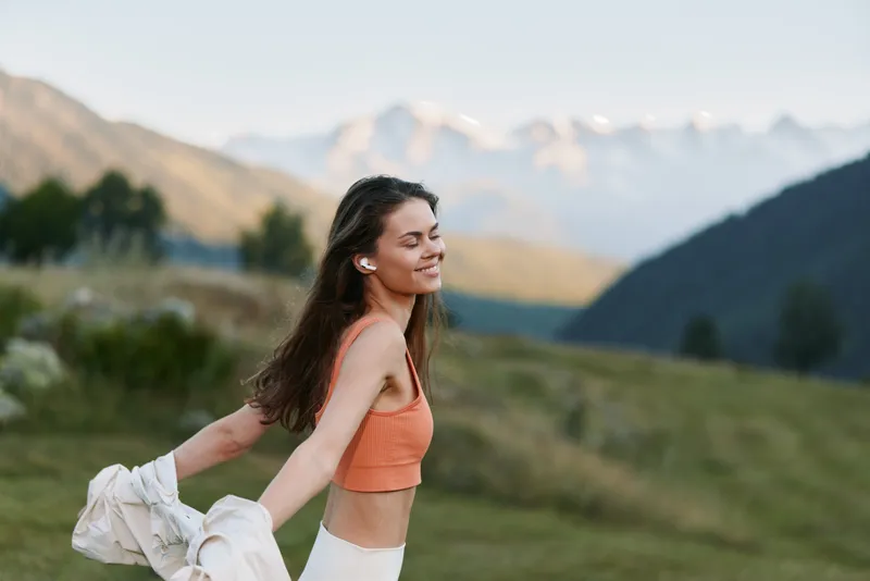 A cheerful woman enjoys an open outdoor moment in a scenic landscape. She wears an orange crop top and smiles, radiating a carefree and vibrant mood.