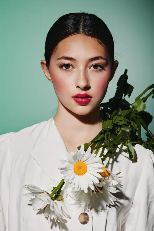 Young woman with creative bright makeup and red lips poses holding white daisies on her bare shoulder on green background. Concept for beauty fashion and creativity.
