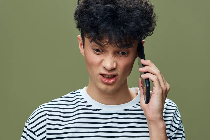 frustrated young man talking phone, curly hair, striped shirt, isolated green background, emotion of anger and confusion, casual style