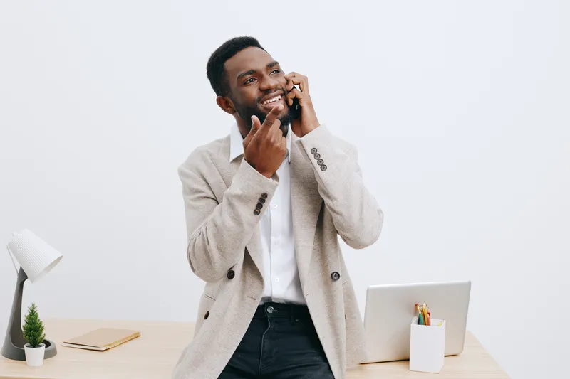 Friendly smiling young African American man talking on mobile phone and laughing while standing in modern office interior with desk and laptop, expressing happiness and positive mood. People lifestyle