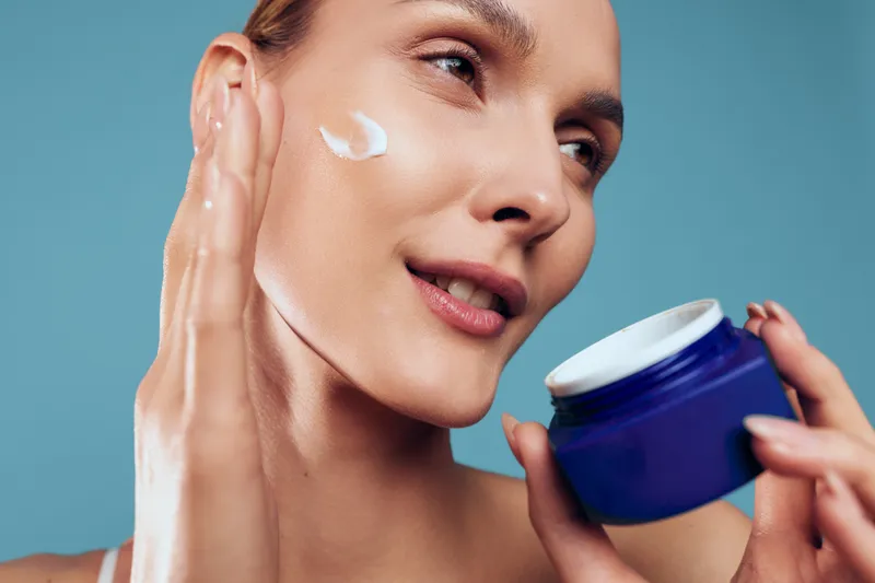 Skincare woman face moisturizer cream blue jar spa beauty routine, female applies cream on cheek, holds blue jar of moisturizer, smooth skin, portrait, blue backdrop