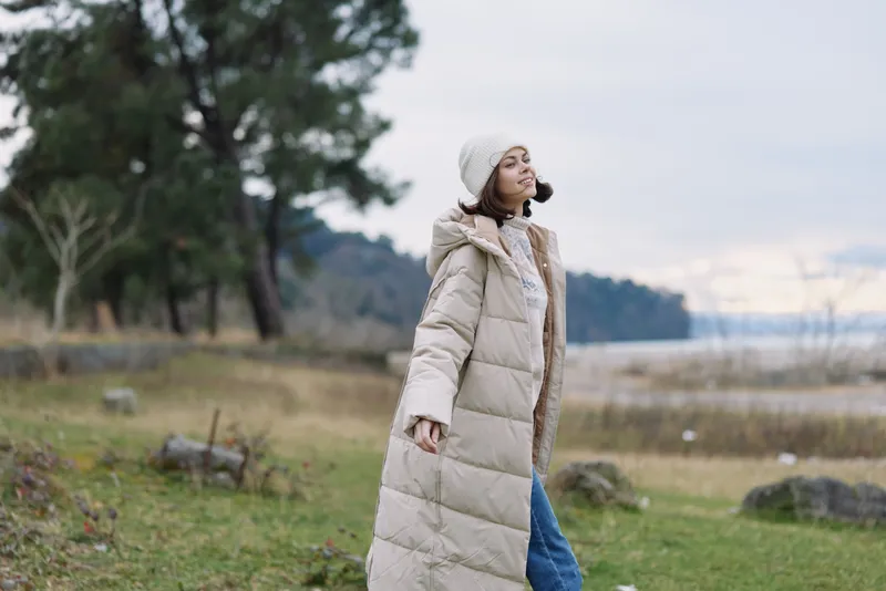 Outdoor woman in a long beige winter coat strolling by a lake in a tranquil park, capturing calm scenery and breezy attitude against distant mountains and open sky.