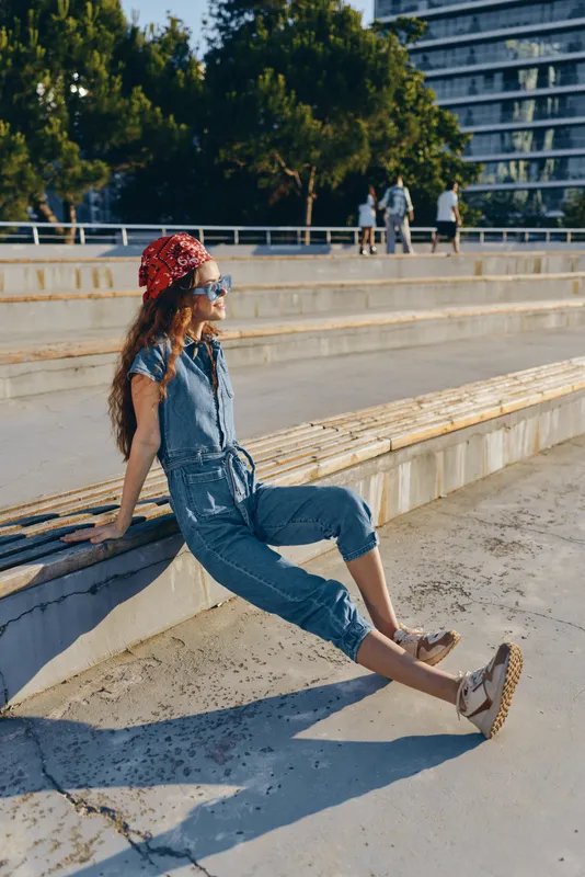 relaxed woman in denim jumpsuit and red bandana sitting outdoors by concrete steps on a sunny day with trees and urban buildings in the background