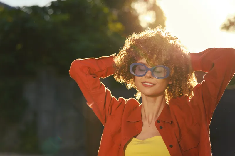 Smiling young woman with curly hair poses outdoors in a bright orange jacket and yellow top, sunglasses perched, as warm backlight creates a sunny glow