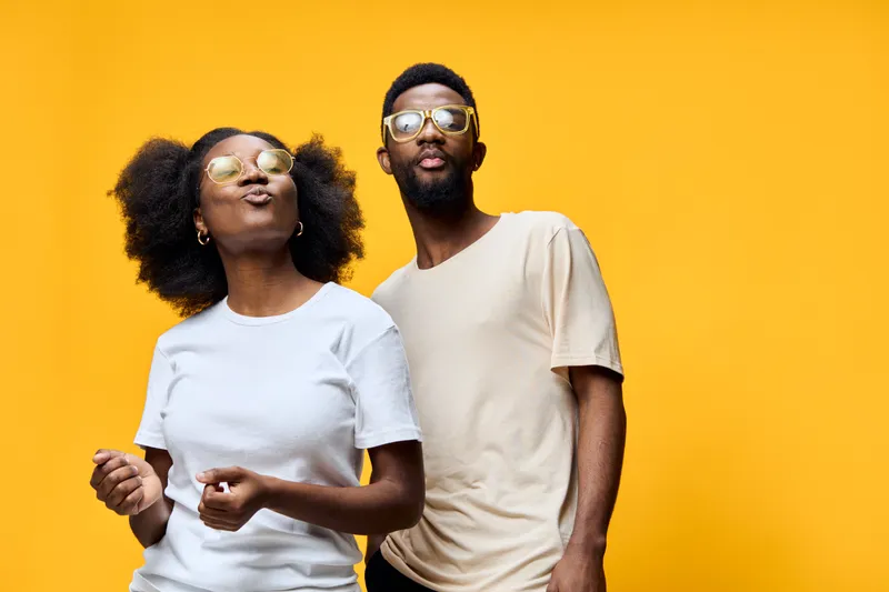 Joyful young African couple in casual outfits, enjoying life against a vibrant yellow background, exuding confidence and happiness with stylish sunglasses