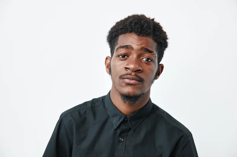 Young african man with dark skin and short curly hair wearing black shirt looking at camera with neutral expression on white background.