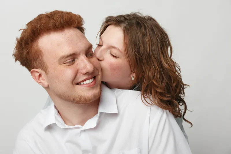 Young couple in white shirts share a tender moment as woman kisses man on cheek. Close-up on light background. Concept of love, affection, relationship, happiness, and intimacy.