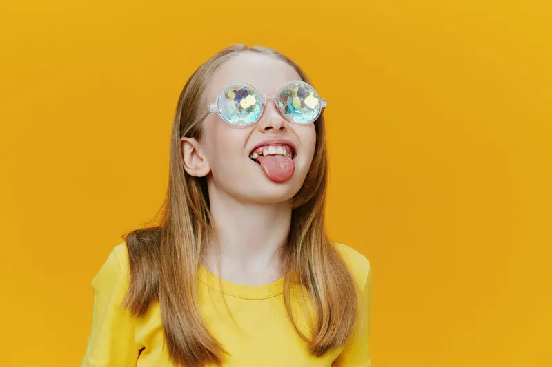 A playful young girl with sunglasses sticking out her tongue, wearing a bright yellow t shirt against a vibrant yellow background, evoking joy and fun in summer