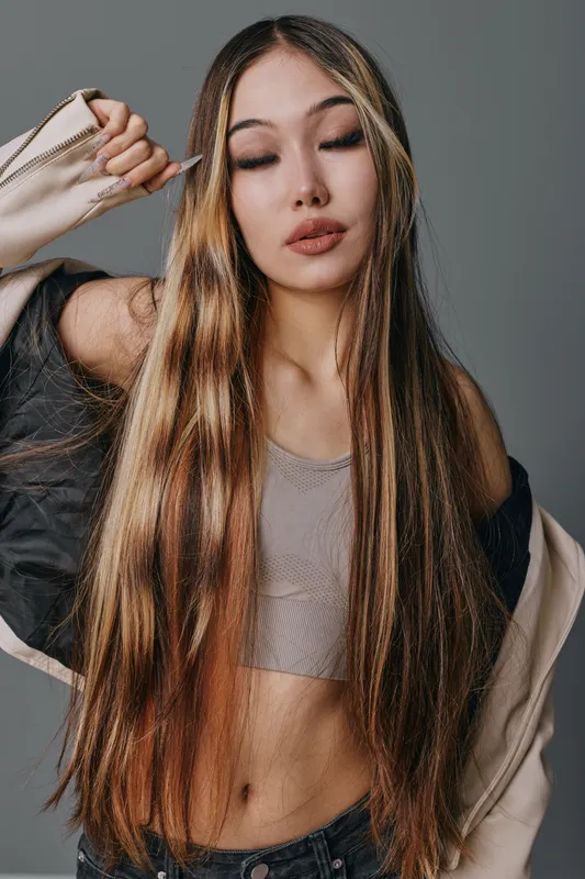 Young woman with long straight hair poses with eyes closed wearing casual top and jacket in studio. Fashion model shows confident relaxed expression with natural makeup and trendy style.