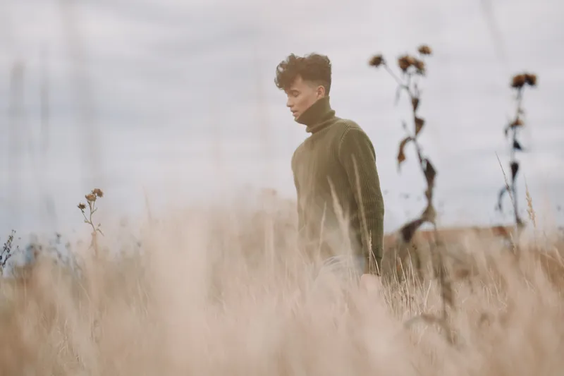 A contemplative young man walks through tall dry grass in muted light, captured with natural tones and genuine emotion to convey authenticity and quiet credibility in a candid outdoor portrait.