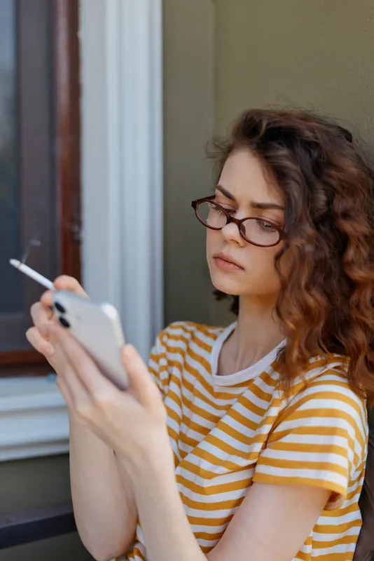 Woman with glasses in a yellow striped t shirt sits on a porch, looking at her smartphone, holding a cigarette, capturing a casual lifestyle moment outdoors.