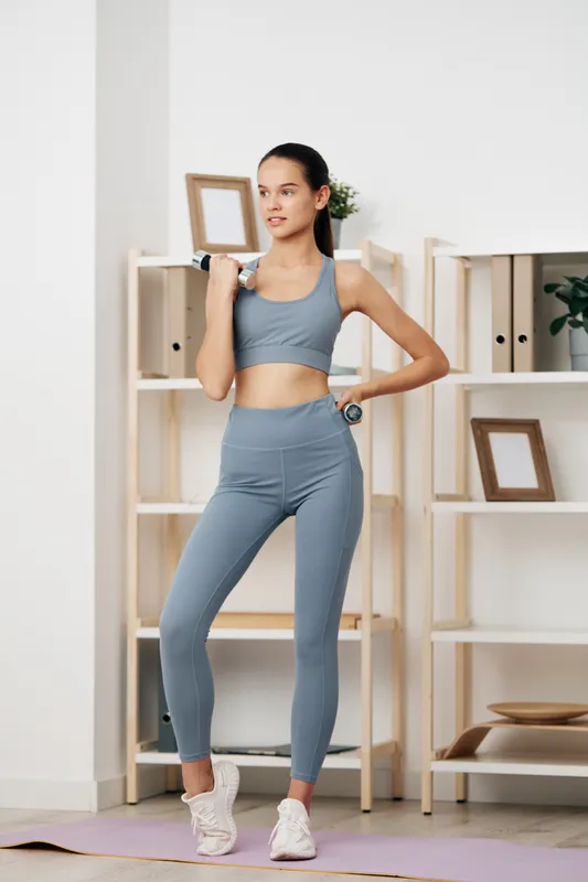 Female athlete in light blue fitness wear performs a dumbbell squat with focused posture in a clean home gym, neutral background, minimal shelves, and natural light.