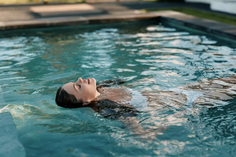 Young woman floating on her back in an outdoor pool with eyes closed, enjoying a peaceful moment of relaxation and lifestyle in soft natural light.