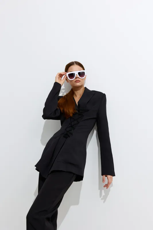 Fashion woman in a stylish black outfit and white sunglasses posing confidently against a bright white background, showcasing contemporary beauty and style.