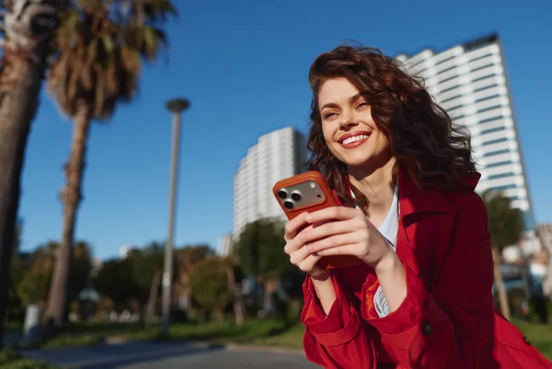 Smiling woman in red coat holding smartphone outdoors, sunny day, modern city background, casual style, natural light, happy expression, urban park setting