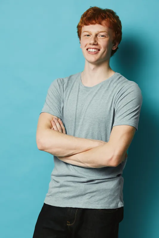 Young man with red hair in gray t shirt standing with crossed arms smiling on blue background casual confident portrait of male model with freckled face and relaxed pose