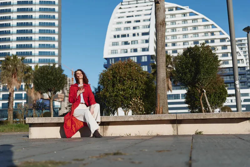 Young woman with curly hair wearing a red jacket and white pants sits on a concrete ledge in a sunlit urban plaza, smiling softly as modern buildings loom behind.