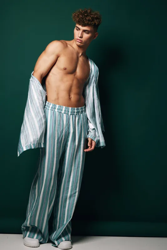 Young man with curly hair posing shirtless wearing striped loose pants and open shirt against dark background. Fashion model with toned body and casual style in studio lighting.