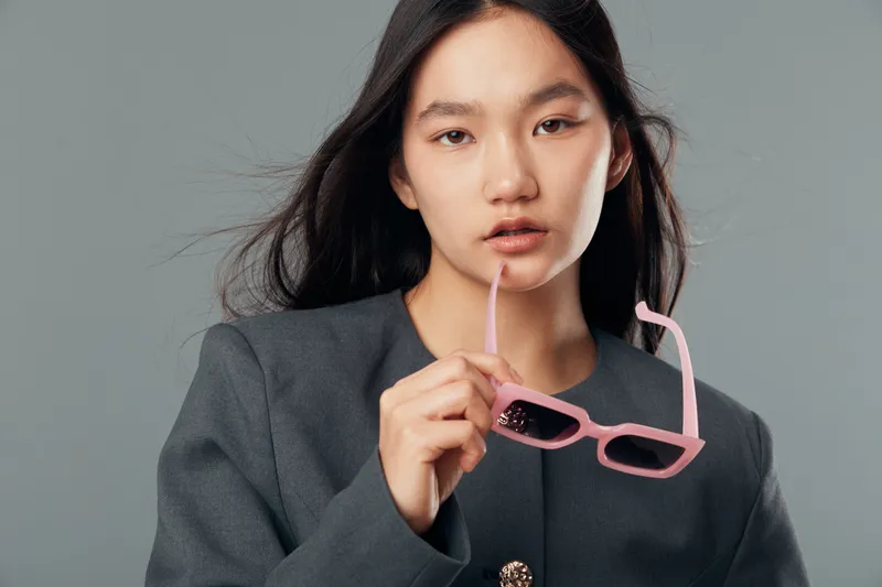 Woman with long dark hair holds pink sunglasses near lips wearing gray jacket on gray background with serious expression and natural makeup for fashion portrait.