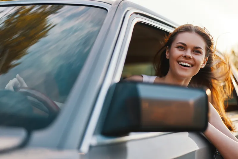 Cheerful young woman enjoying a road trip, leaning out of a vintage car window with flowing hair and a bright smile, captured in the golden hour light.