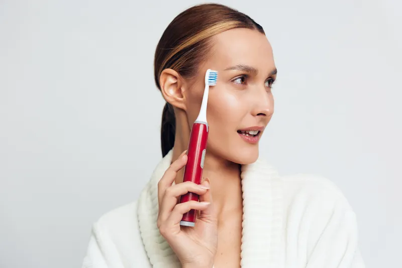 A woman in a white robe holds a toothbrush near her temple, examining her oral care routine in a bright, clean setting. This scene emphasizes personal grooming and wellness through daily care.