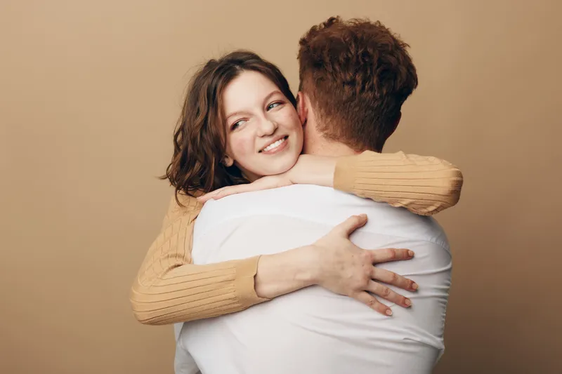 Smiling woman with brown hair hugs man wearing white shirt on beige background. Intimate moment of affection and connection between couple. Concept of love, relationship, and happiness.