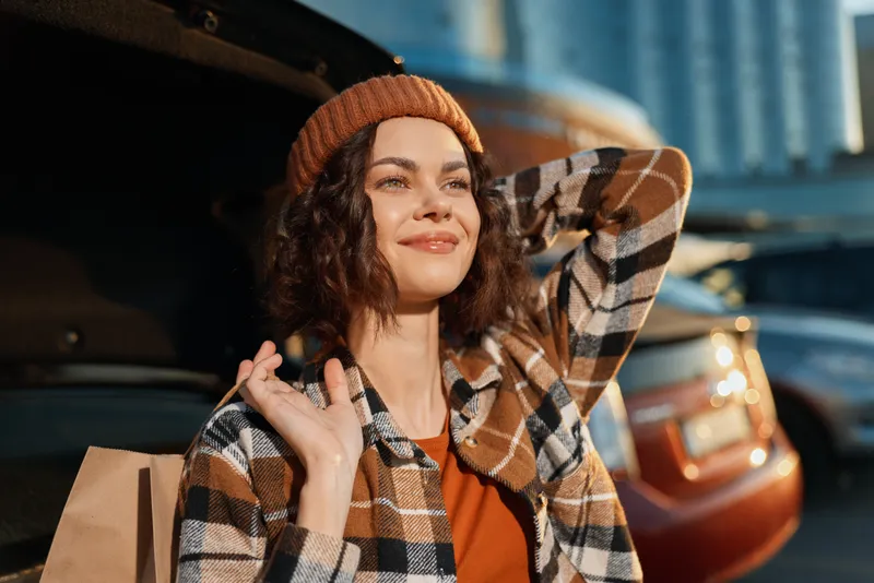 Woman smile by car trunk after shopping in urban setting, wearing beanie and plaid coat with golden hour glow, candid lifestyle portrait emphasizing authenticity, mindful living and emotional