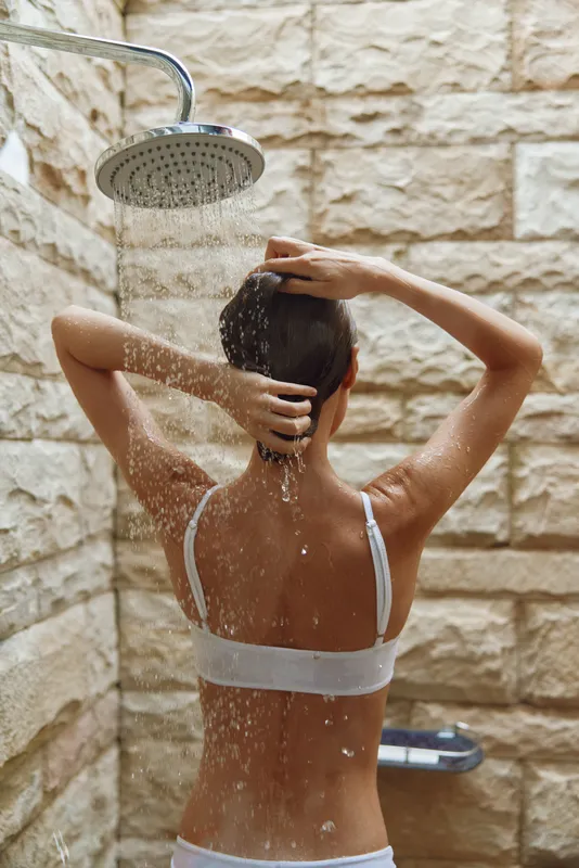 woman showering outdoors under rain showerhead with stone wall background, natural light, health and hygiene concept in warm earth tones