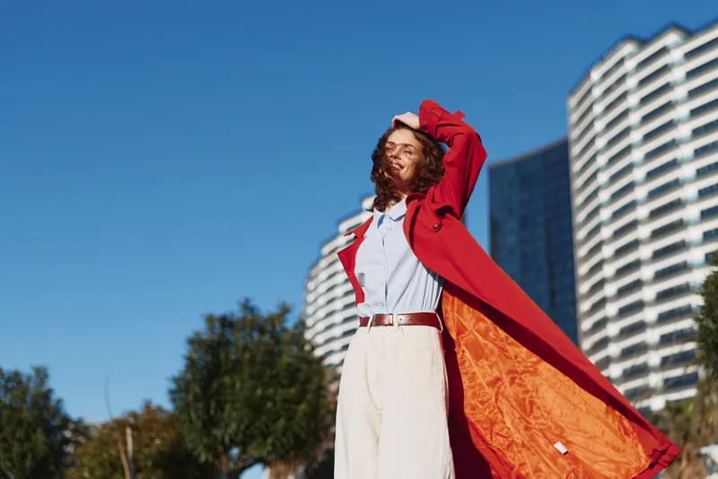 Young woman in a red coat enjoys a sunny day outdoors while standing confidently with eyes closed and hand on her head.