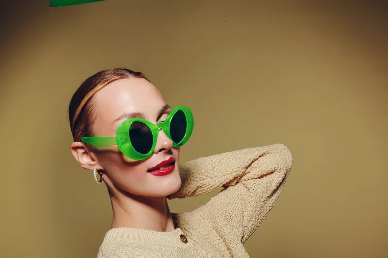 Stylish woman with green round sunglasses and red lipstick poses confidently against beige background. Fashion concept with trendy accessories and casual knitwear.