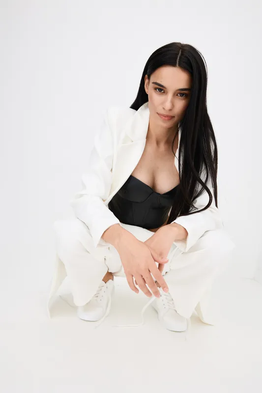Stylish woman crouching in white suit and black corset top with long dark hair and clean minimalist background, conveying modern fashion and confidence.