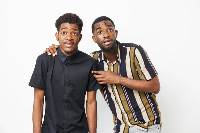 Two surprised African American men express excitement and curiosity, showcasing unique styles and emotions against a clean background.