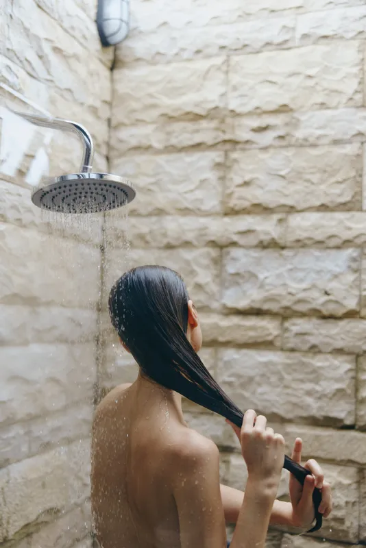woman showering outdoors under running water with wet hair against rustic stone wall, natural light, relaxing moment, refreshing bathing experience