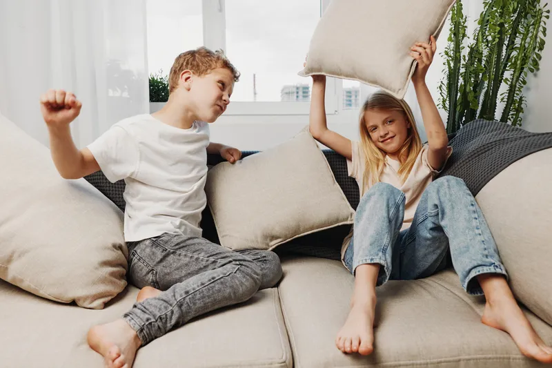 Two playful children having a pillow fight on a beige couch in a bright living room, smiling and enjoying a fun moment together.