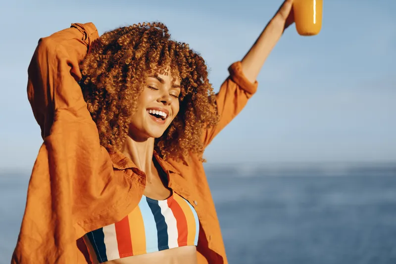 Curly haired woman laughing outdoors at seaside in summer orange jacket striped shirt arms raised golden light ocean horizon blue sky joyful smile celebrating freedom casual lifestyle