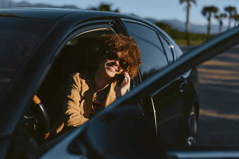A young woman with curly hair smiles as she sits in a car window during a casual drive, conveying a relaxed, confident moment of everyday life.