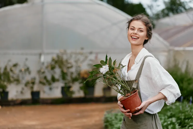 Happy young woman holding a potted plant in a greenhouse, dressed casually in a white blouse and overalls, surrounded by greenery and soft natural lighting