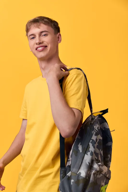 Youth wearing a yellow shirt with a camo backpack poses against a bright yellow backdrop, exuding casual style and travel ready vibe for lifestyle, education, and everyday activity themes